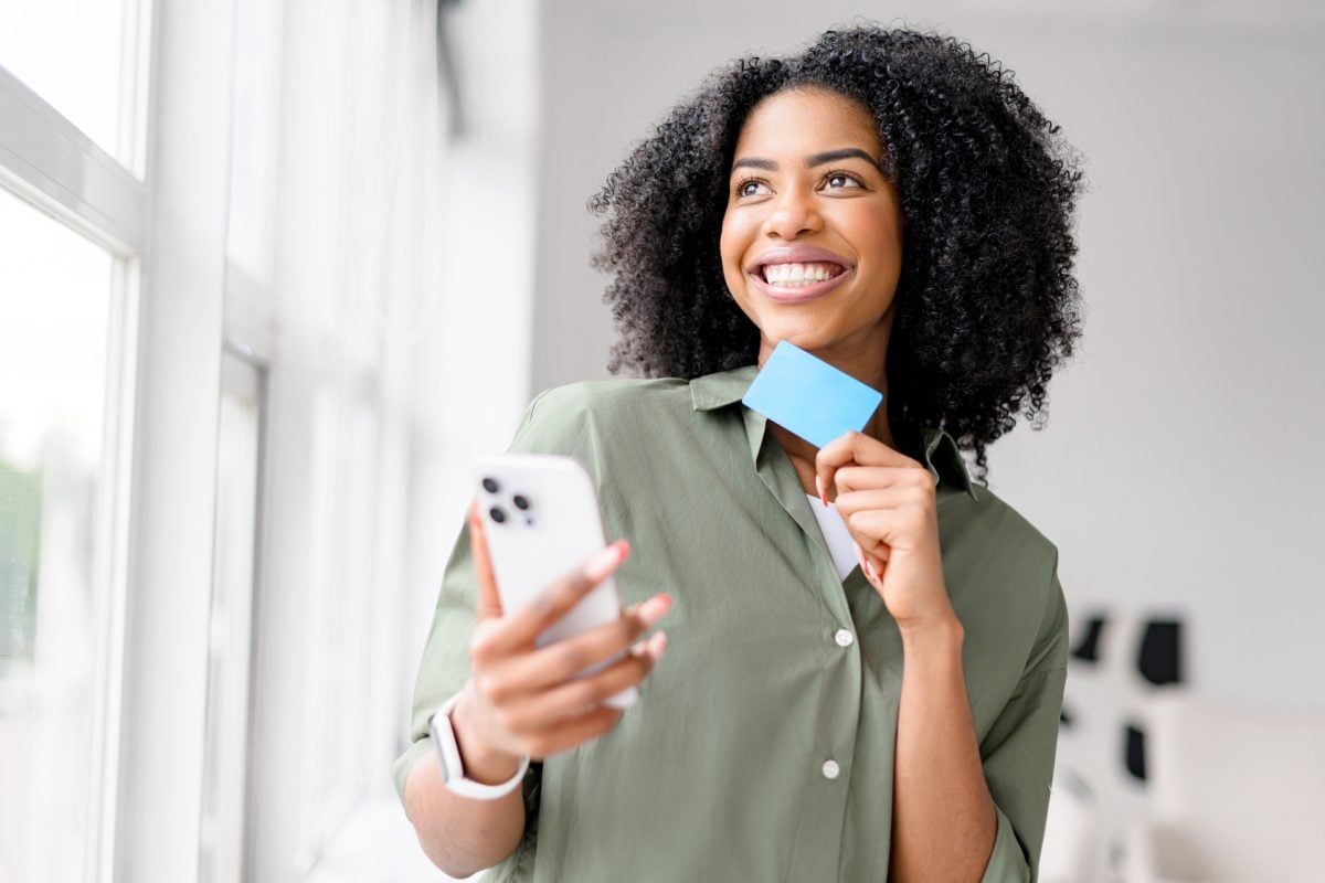 An African-American woman laughs lightly while looking at her smartphone, a blue credit card in hand suggesting a joyful online shopping experience or the ease of managing finances digitally. 